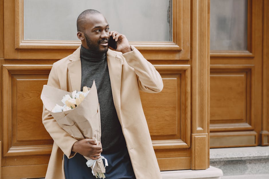 A man with dark skin and a beard, dressed in a beige overcoat and dark gray turtleneck, is standing outdoors in front of a wooden-paneled building. He is holding a bouquet of fresh white and cream flo